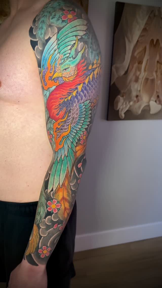 Colorful phoenix sleeve tattoo on a man's arm with clouds and flowers.