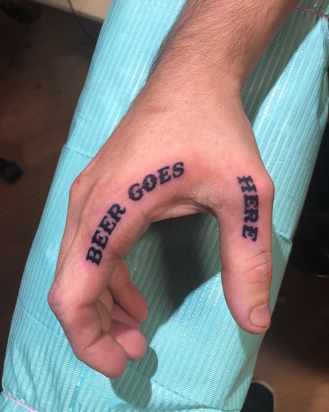 Hand with black script tattoos reading BEER GOES HERE across knuckles and thumb.