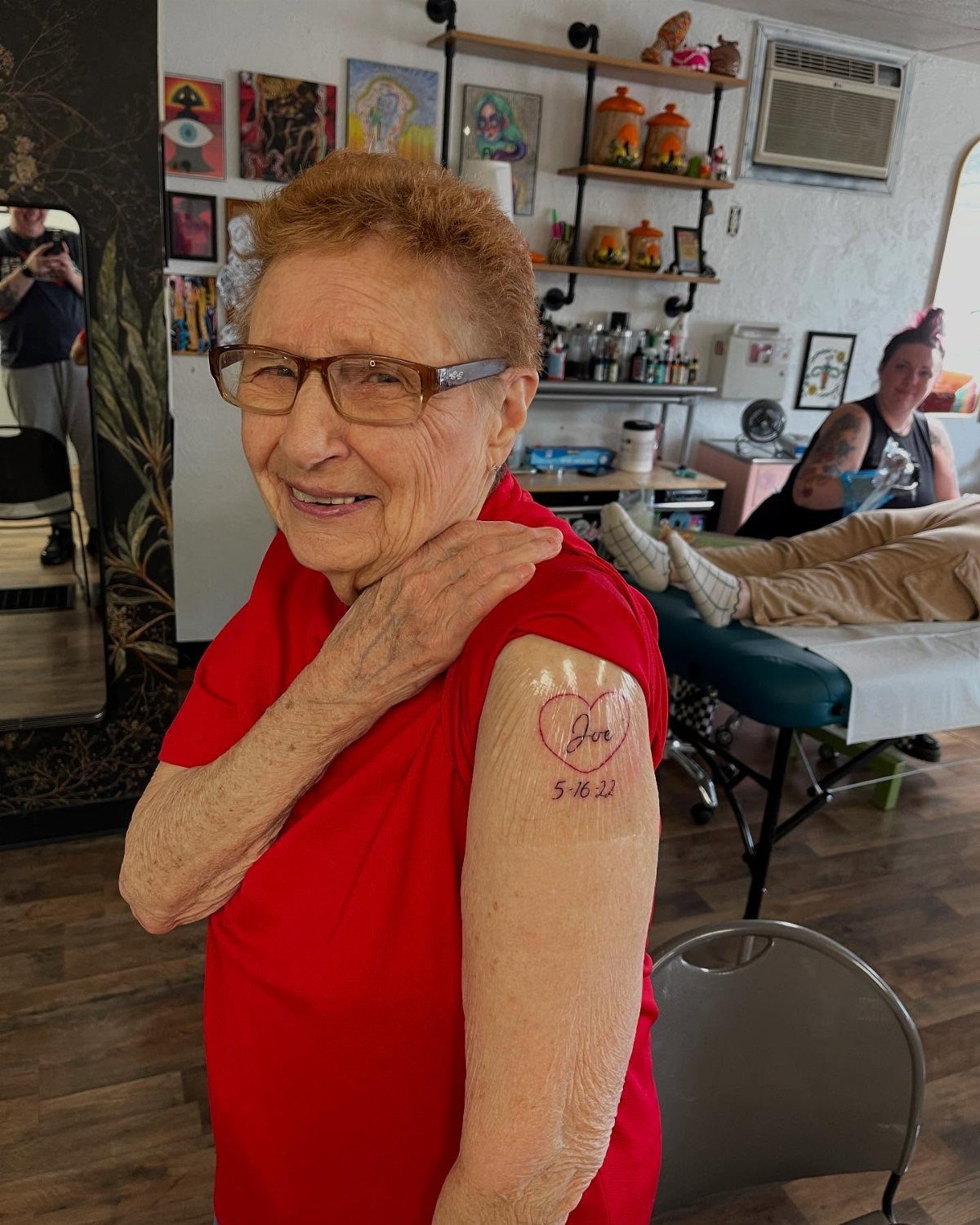 Older woman in red shirt showing a heart tattoo with a name on her arm.