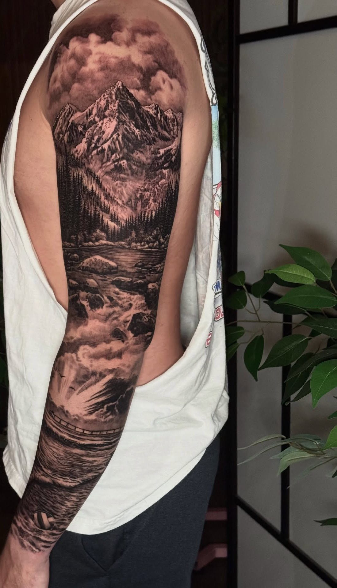 Detailed black and grey sleeve tattoo of mountains, pine trees, and a river.