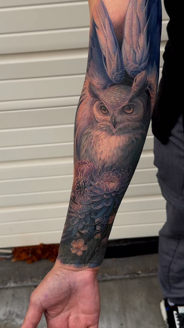 Realistic owl tattoo on a man's arm with floral details and blue shading.