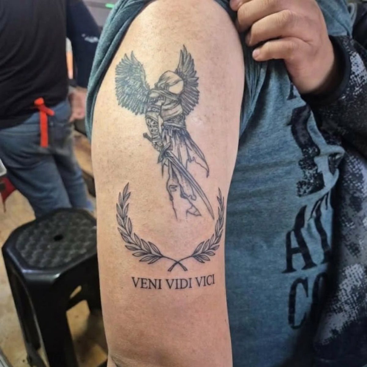 Man's arm tattoo featuring a winged figure, laurel wreath, and Veni Vidi Vici text.
