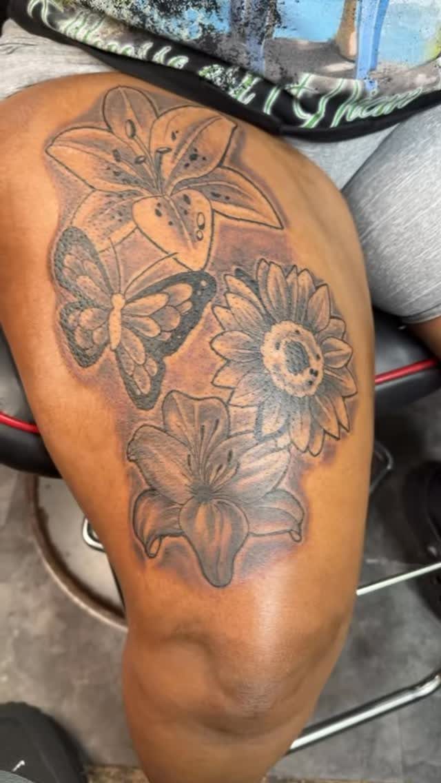 Thigh tattoo featuring a butterfly combined with lilies and a sunflower in grey ink.