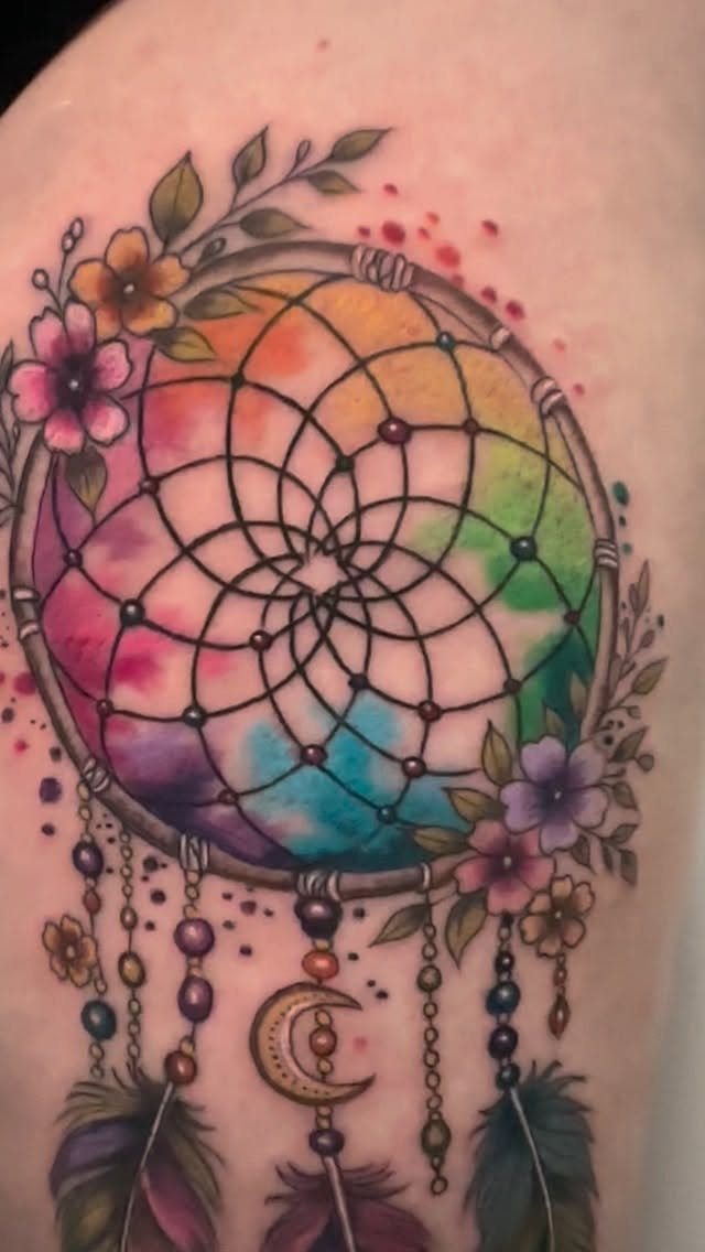 Colorful dreamcatcher tattoo with rainbow watercolor and feathers on a woman's thigh.