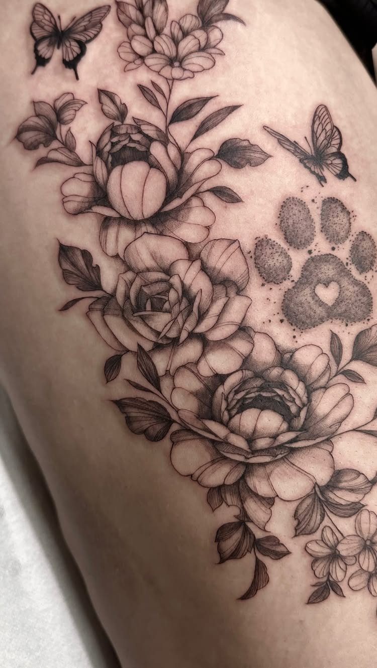 Floral thigh tattoo with peonies, roses, butterflies, and a paw print in black ink.