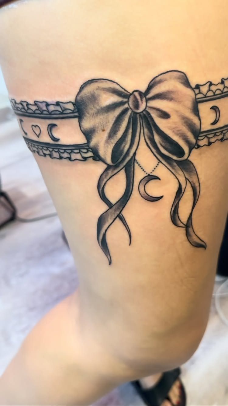 Lace garter thigh tattoo with a large bow and crescent moon charm.