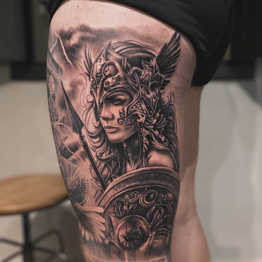Female gladiator tattoo with helmet and shield on a woman's thigh in black and grey.
