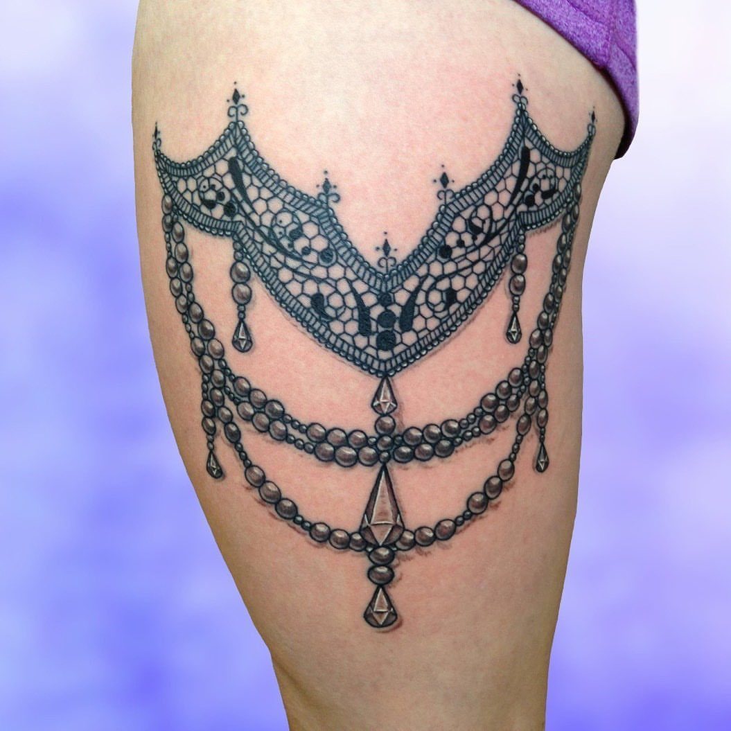 Lace tattoo on a thigh featuring intricate patterns, beads, and diamond charms.