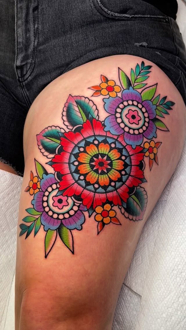 Colorful floral and mandala tattoo on a woman's thigh with red and purple flowers.