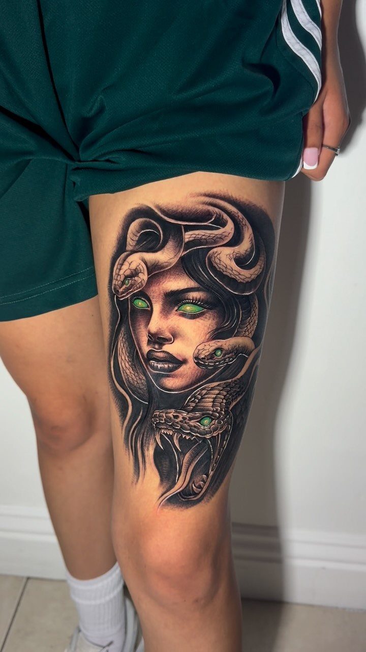 Medusa tattoo on a woman's thigh with snakes for hair and green eyes