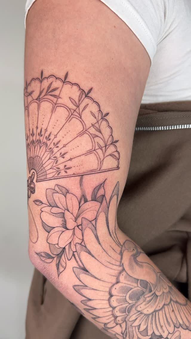 Tattooed arm showing a traditional folding fan, a flower, and a bird in black ink.