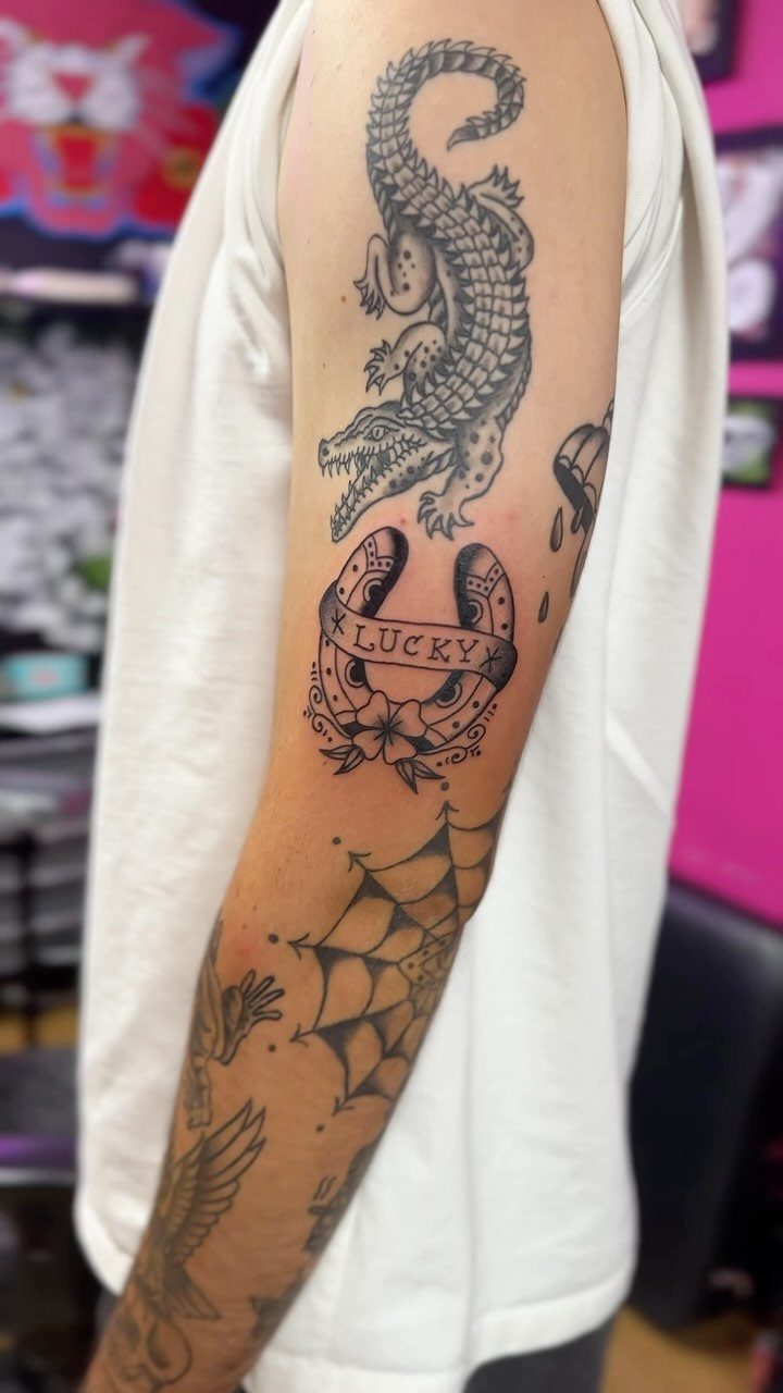 Tattooed arm showing a lucky horseshoe with flowers and a crocodile above it.