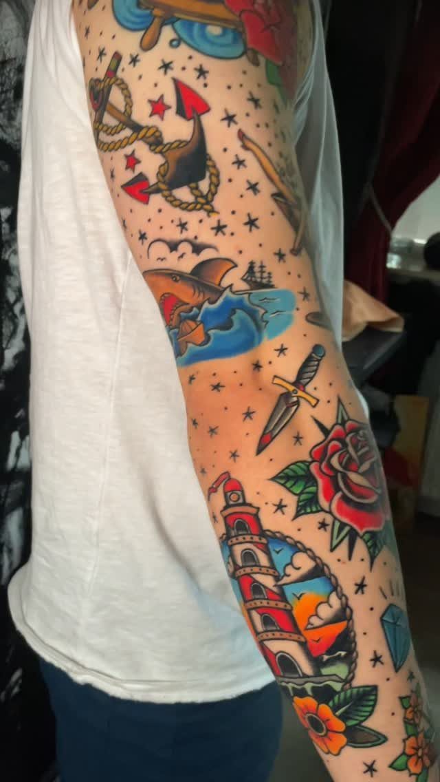 Traditional tattoo sleeve on a man's arm featuring an anchor, shark, and lighthouse.