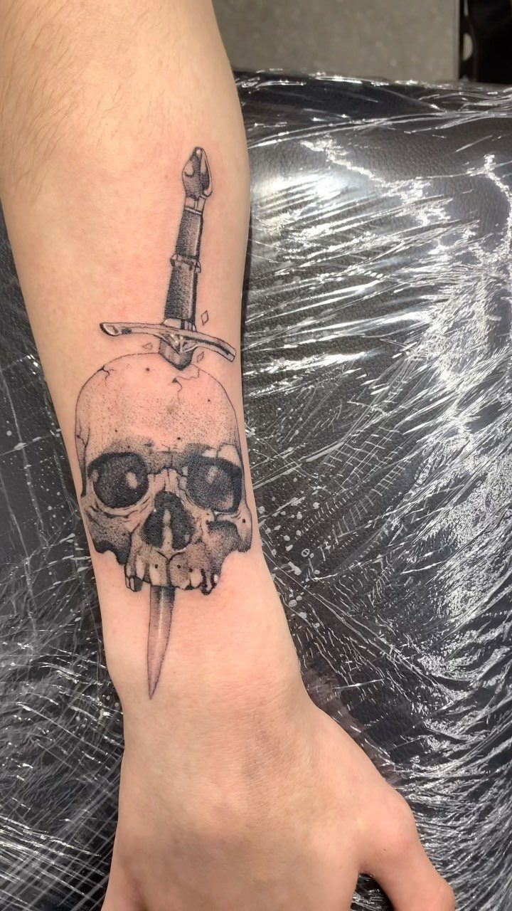 Skull and dagger tattoo on a forearm in black and grey ink.