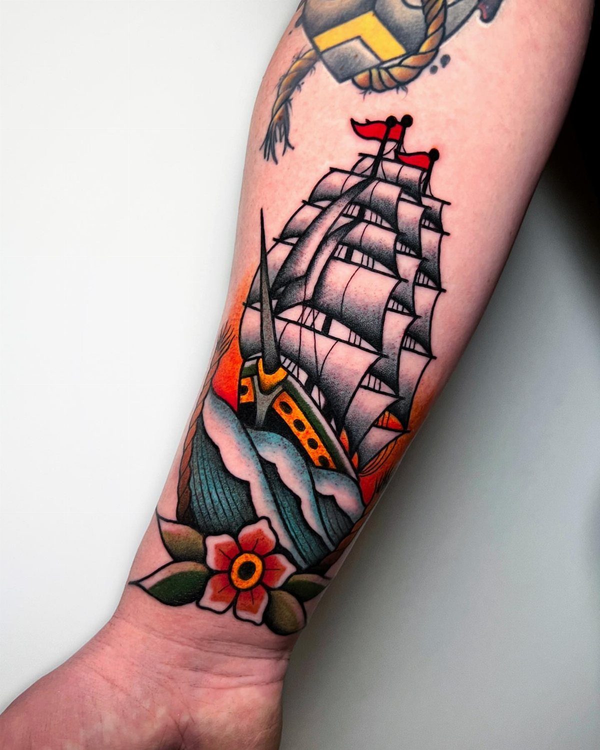 Sailing ship tattoo with white sails and red flags on a forearm.