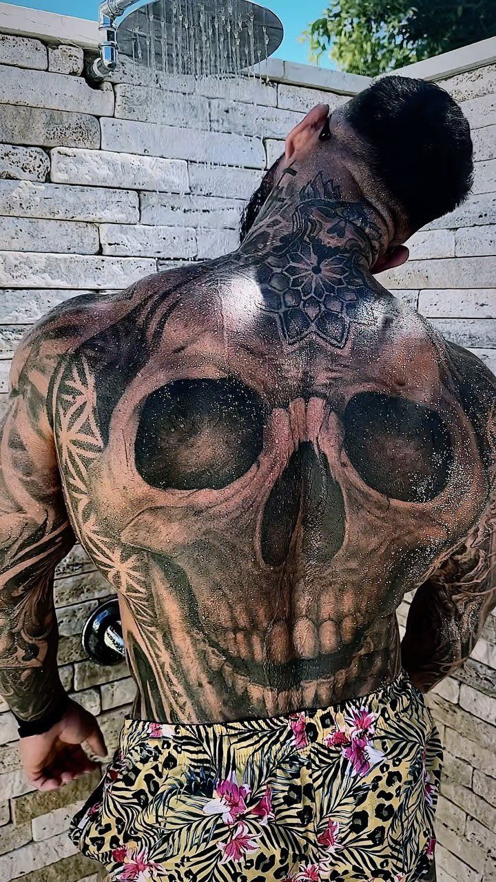Man's back with a large, detailed skull tattoo and floral swim trunks.