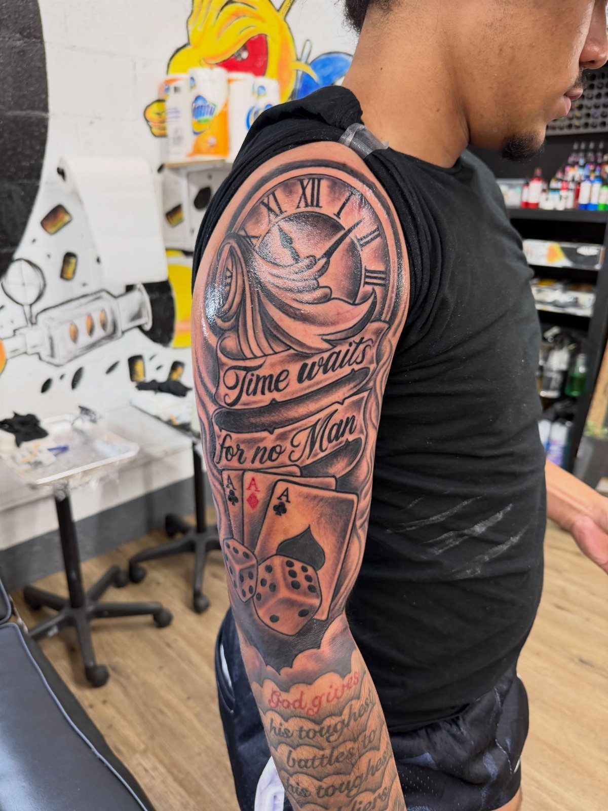 Man's arm tattoo featuring a Roman numeral clock, banner, playing cards, and dice.
