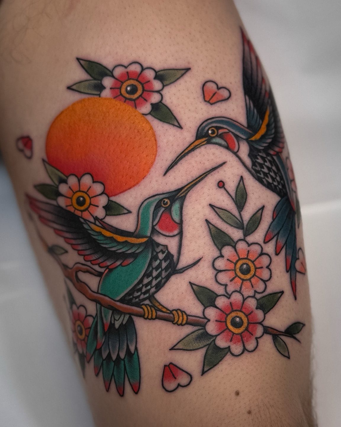 Two traditional hummingbirds with pink flowers and an orange sun tattooed on skin.