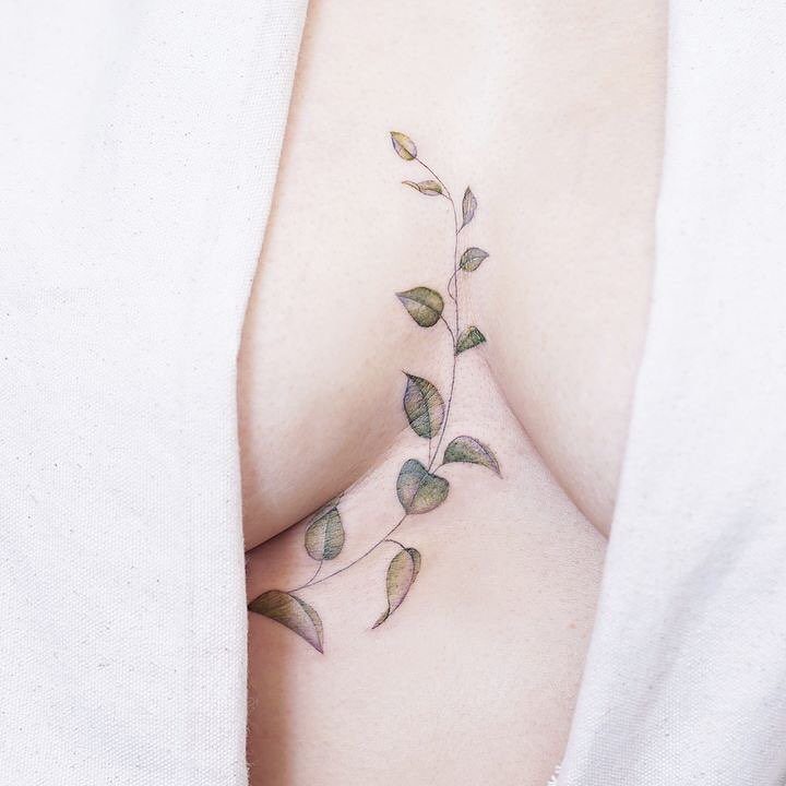 Leafy vine tattoo curving under a woman's breast against white fabric.