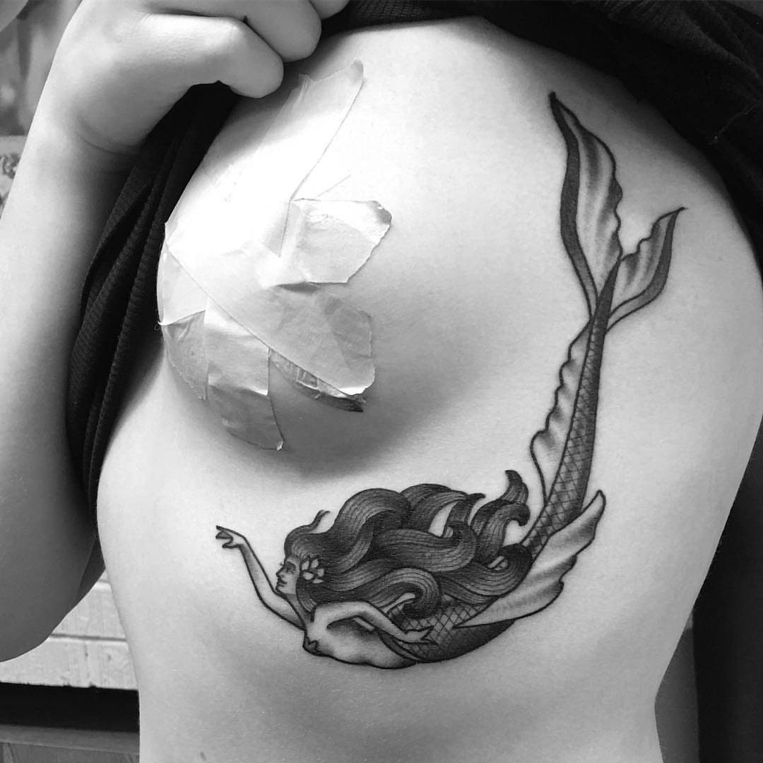 Mermaid tattoo with flowing hair and curved tail on a woman's ribcage.