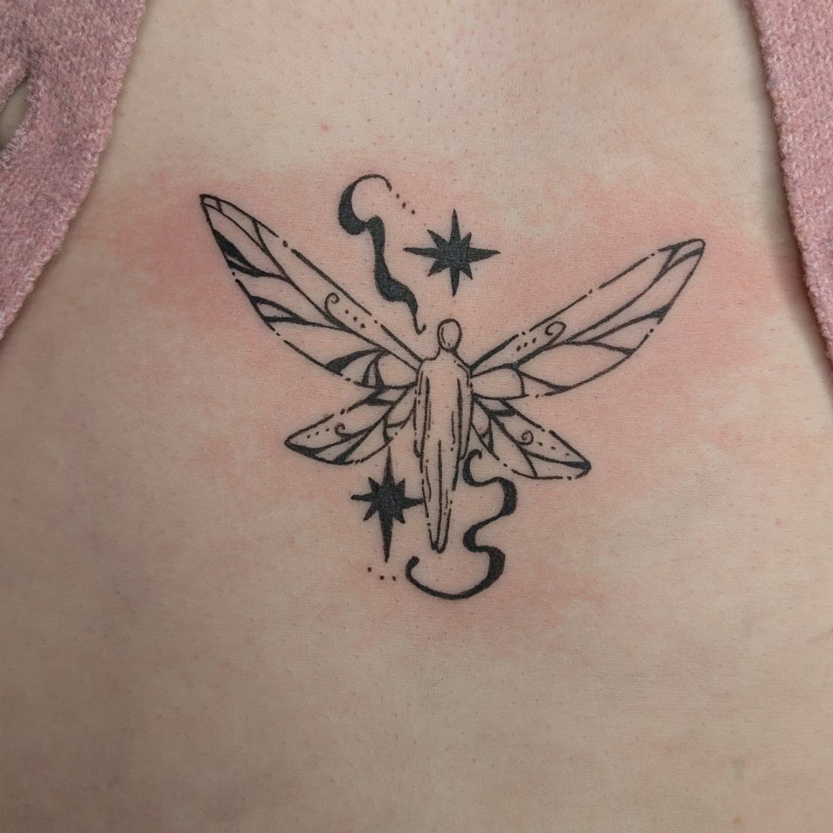 Black ink tattoo of a winged figure and stars located under the chest.