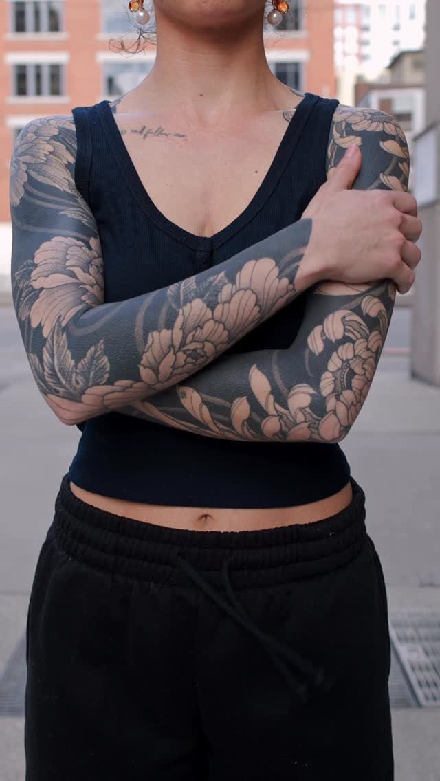Woman with large grey floral tattoos covering both upper arms and forearms.