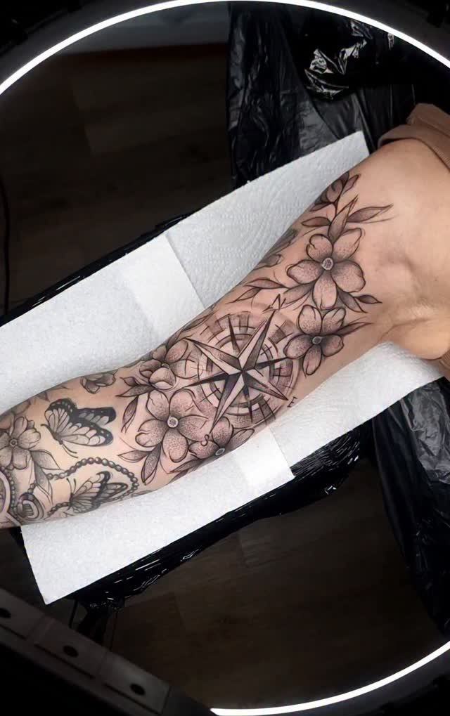 Compass rose tattoo surrounded by flowers and butterflies on a person's upper leg.