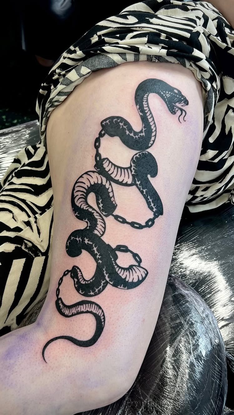 Black ink snake tattoo with a chain on a person's upper thigh.
