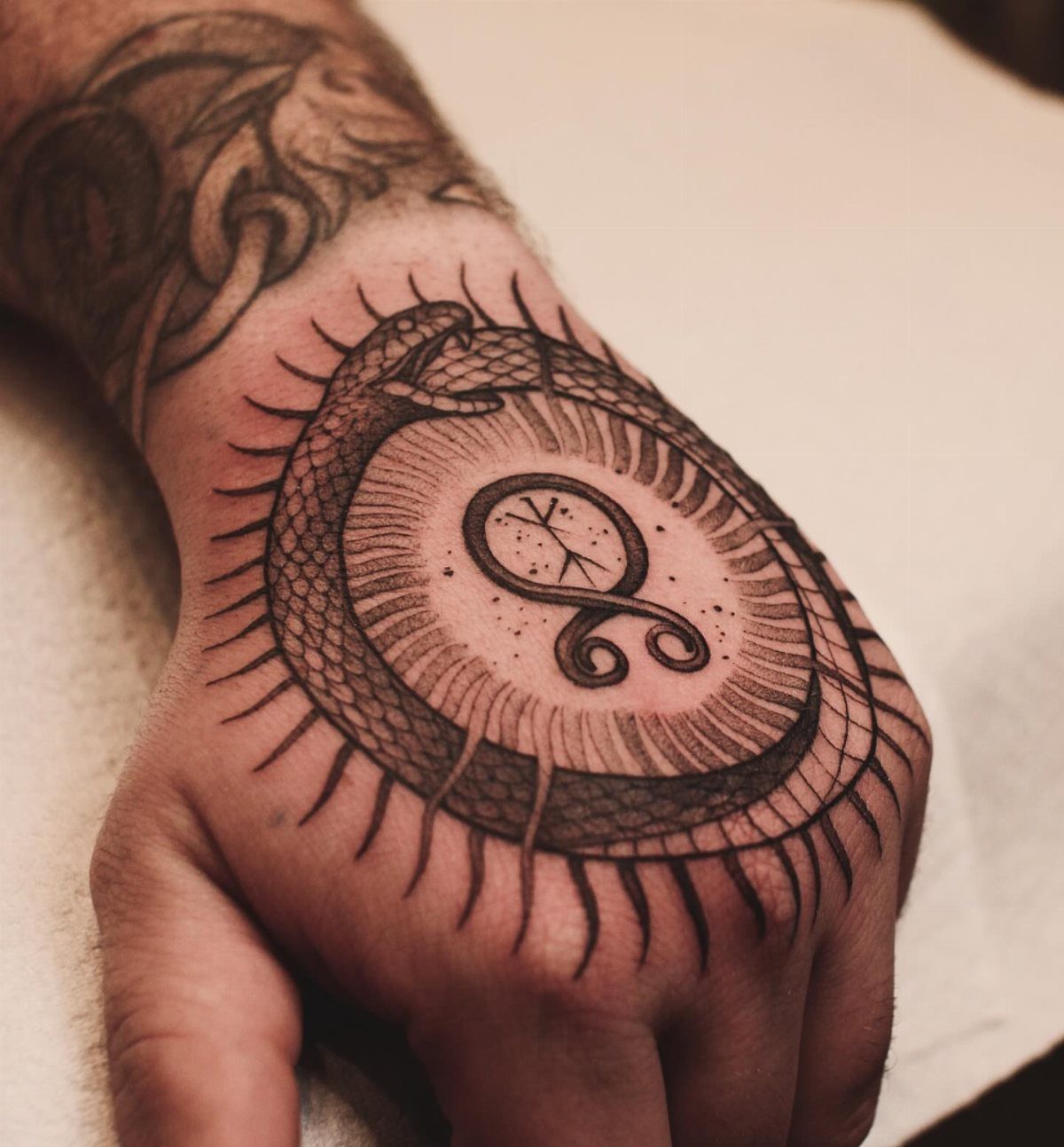 Hand tattoo featuring an Ouroboros serpent encircling a Norse symbol with radiating lines.