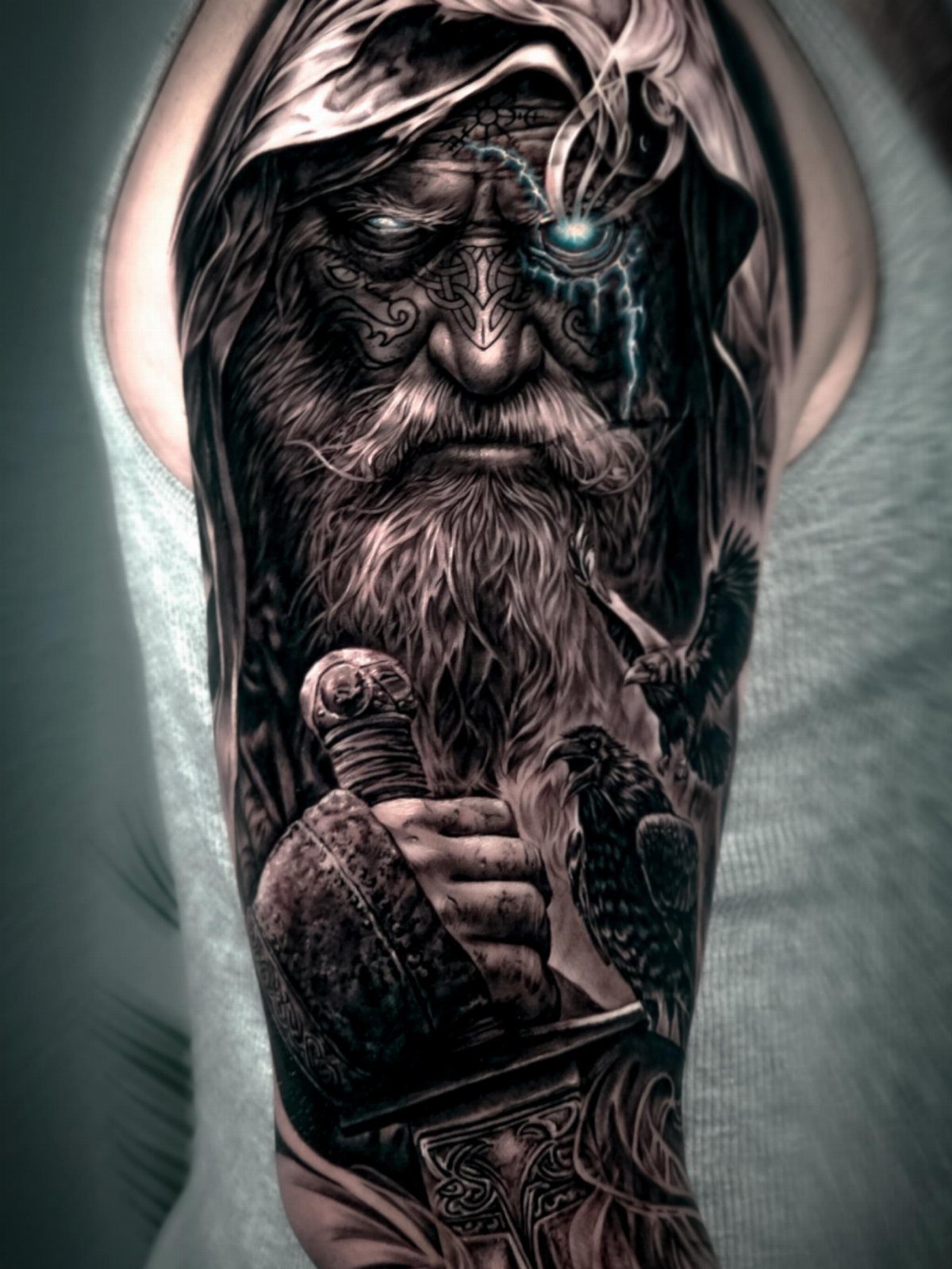 Viking warrior tattoo on arm featuring a hooded man with ravens and a sword.