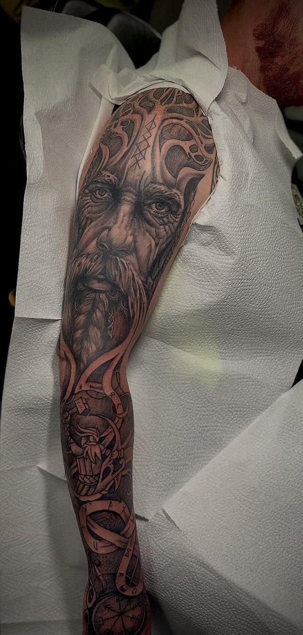 Viking face and Norse symbols tattooed in black and grey ink on an arm.