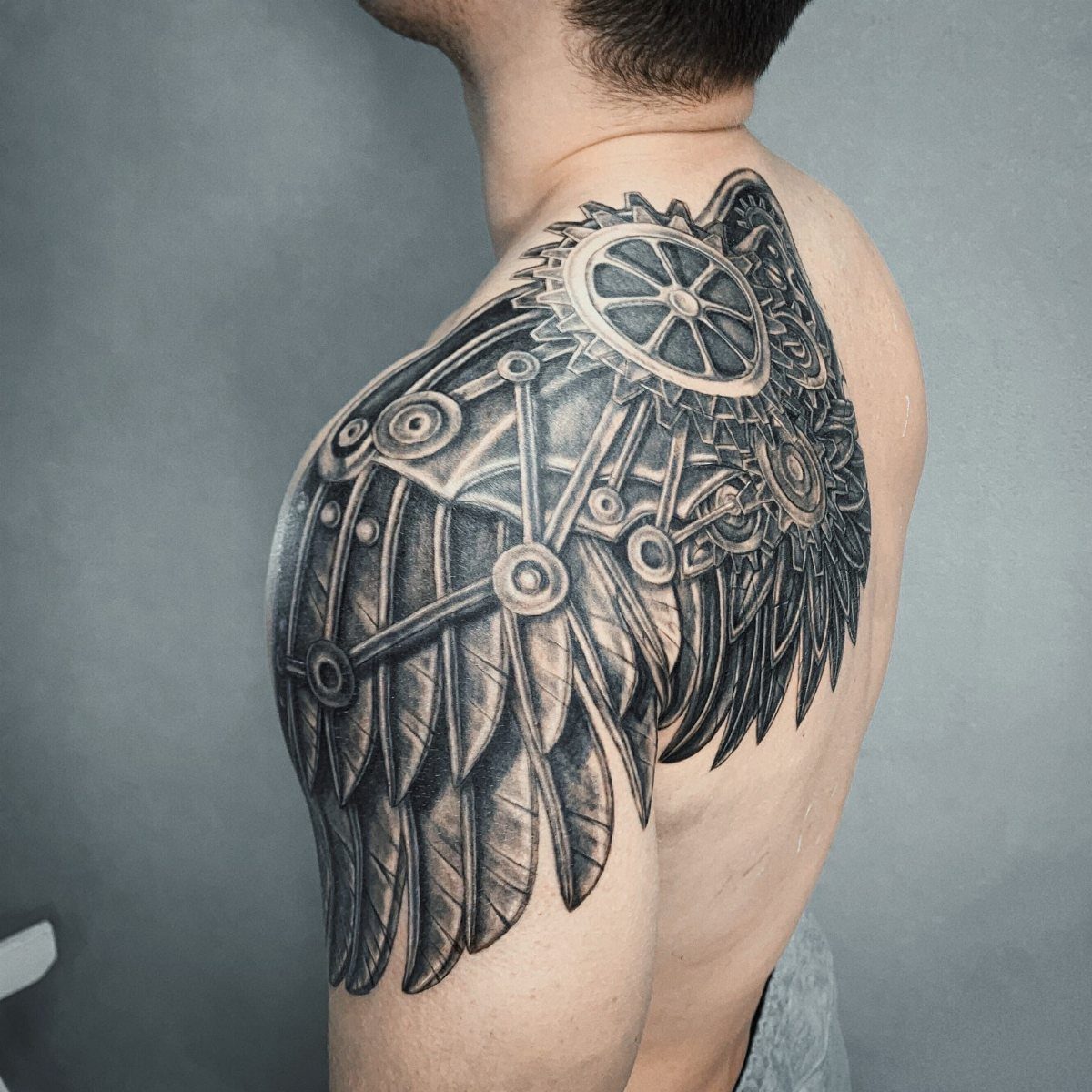 Mechanical wing tattoo with gears and feathers on a man's upper back and shoulder.