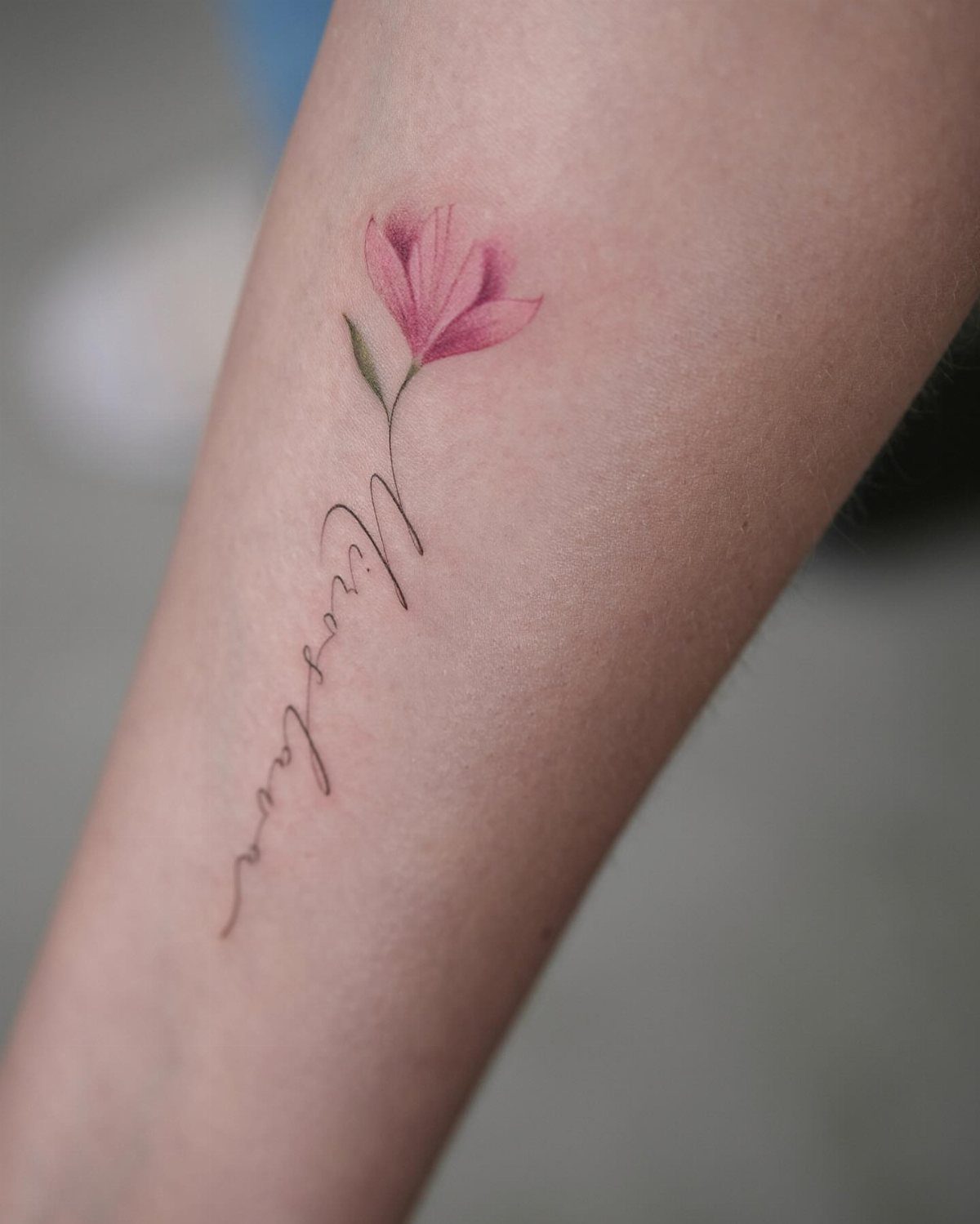 Tattoo of a pink flower with a cursive name written on a person's skin.