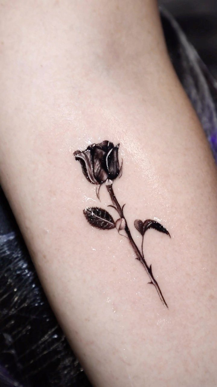 Small black rose tattoo with stem and leaves on fair skin.