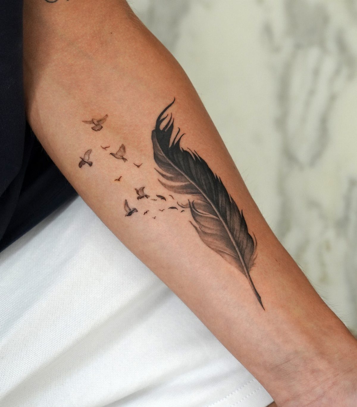 Feather tattoo on a forearm dissolving into a flock of small birds.