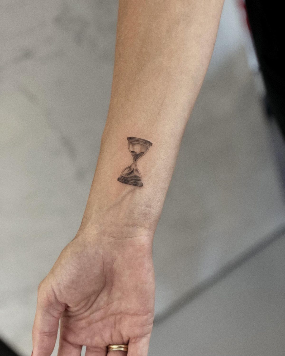 Hourglass tattoo on a person's inner wrist with a gold ring on finger.