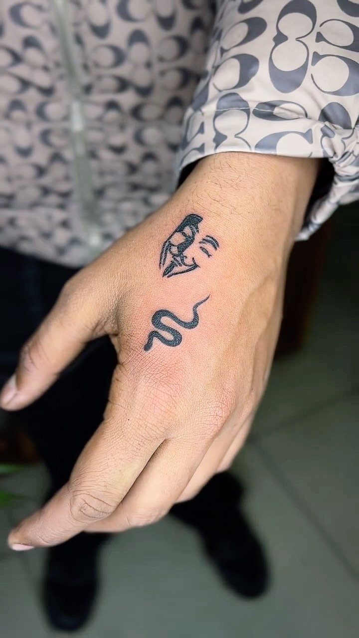 Hand with a small black snake tattoo and a stylized face tattoo.