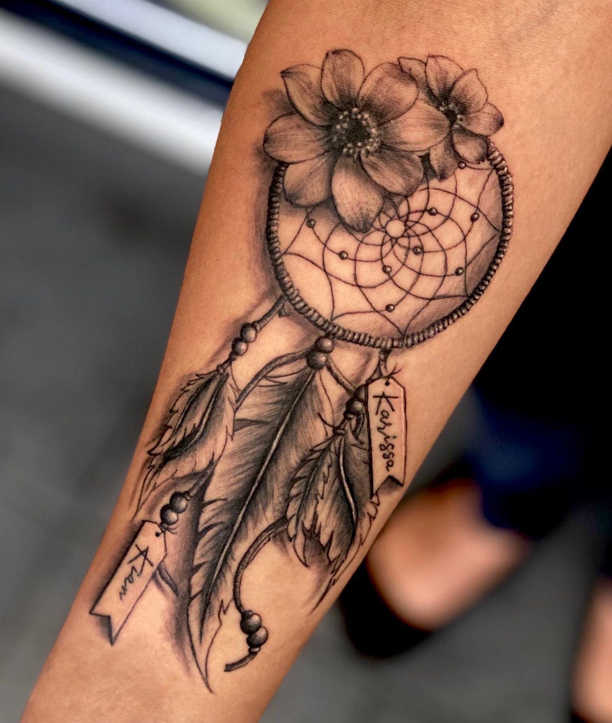 Dreamcatcher tattoo on a forearm with flowers, feathers, and name banners.