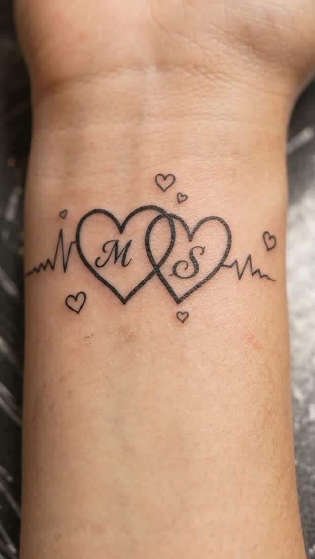 Interlocking heart tattoos with initials on a wrist integrated into a heartbeat line.