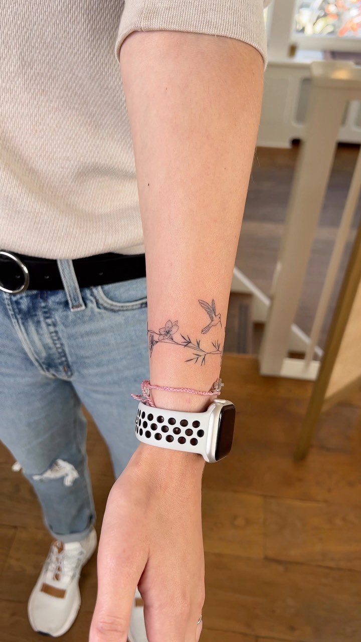 Hummingbird and floral branch tattoo on a person's inner wrist with a smartwatch.