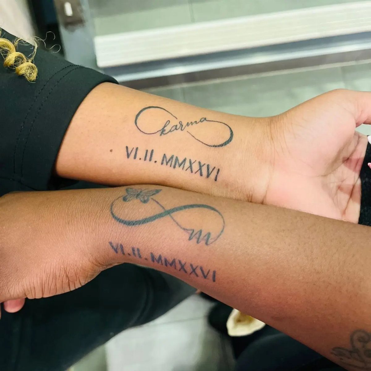 Two wrists with matching infinity symbol tattoos and Roman numerals in black ink.