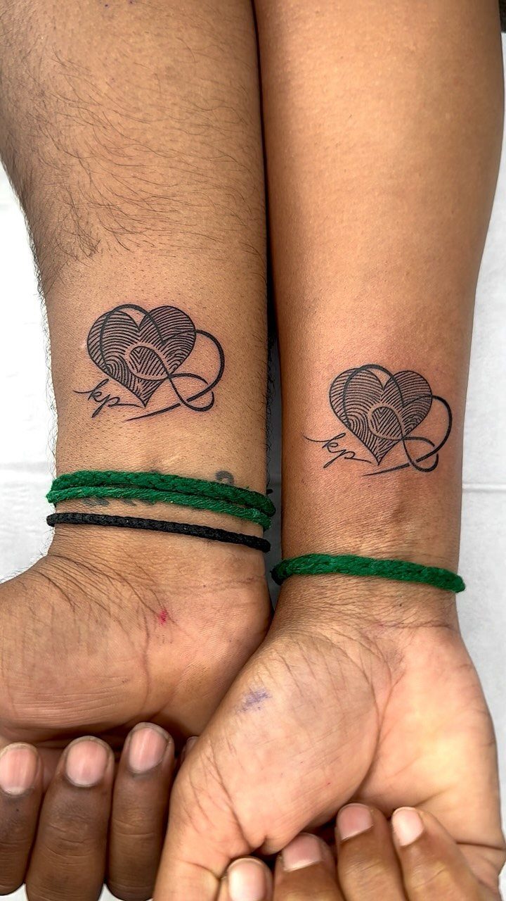 Matching heart and infinity symbol tattoos on two wrists with green bracelets.