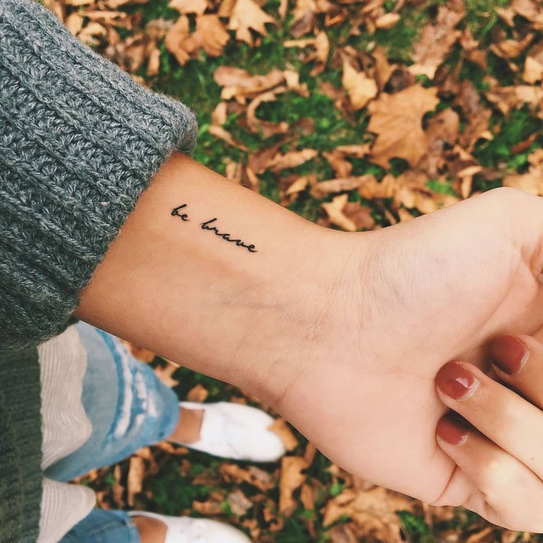Wrist tattoo with the phrase 'be brave' in cursive script on a person's arm.