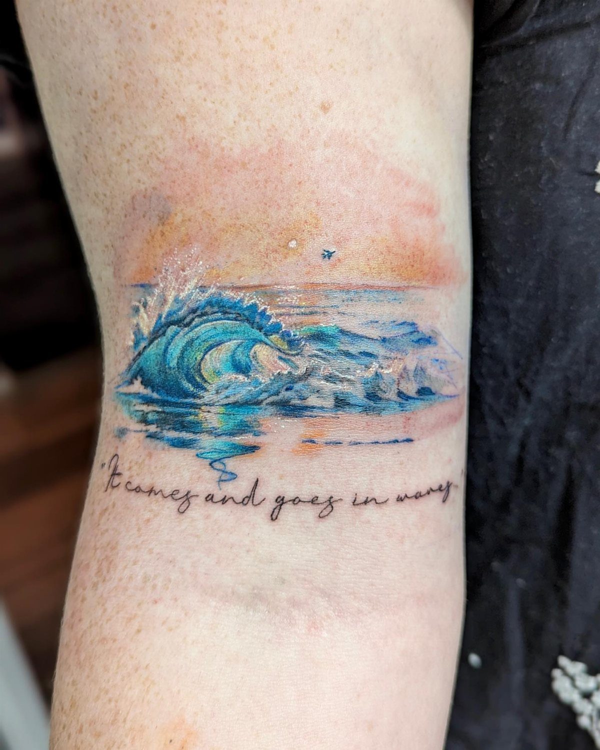 Blue ocean wave tattoo with sunset colors and script on a forearm