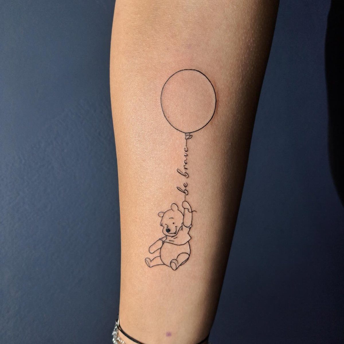 Winnie the Pooh tattoo on a wrist holding a balloon with be brave text