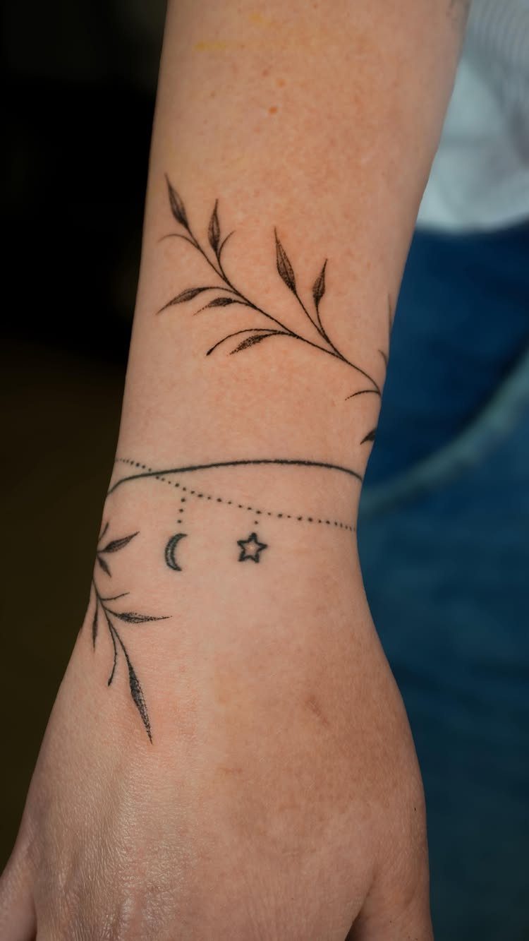 Wrist tattoos featuring a botanical branch and a thin bracelet line with moon and star.
